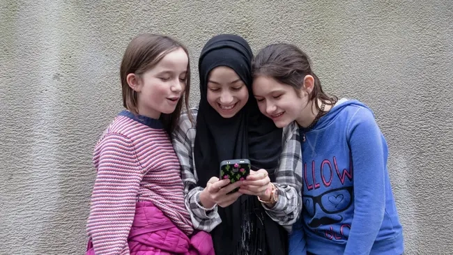 Drei Mädchen stehen nebeneinander vor einer Beton-Wand. Sie alle schauen auf das Handy, das das Mädchen in der Mitte in der Hand hält.