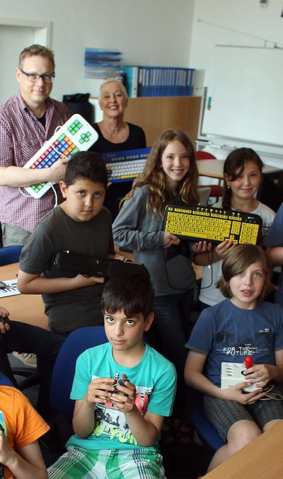 Mehrere Kinder und zwei Erwachsene stehen und sitzen in einem Klassenraum. Jeder von ihnen hält eine assistive Technologie in der Hand.