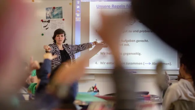 Blick von hinten in eine Klasse: Vorne steht eine Lehrerin an einer digitalen Tafel. Unscharf sind ein paar Arme aufzeigender Kinder zu sehen.