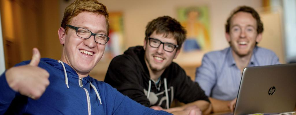 Drei Männer mit und ohne Behinderung sitzen an einem Schreibtisch vor zwei Laptops. Einer von ihnen gibt das Daumen-hoch-Zeichen. Alle drei lachen.