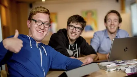 Drei Männer mit und ohne Behinderung sitzen an einem Schreibtisch vor zwei Laptops. Einer von ihnen gibt das Daumen-hoch-Zeichen. Alle drei lachen.