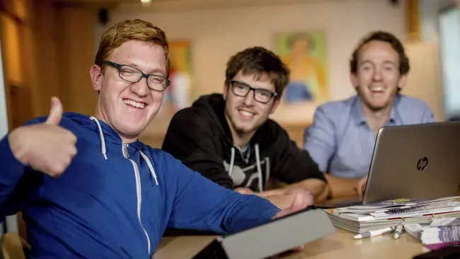 Drei Männer mit und ohne Behinderung sitzen an einem Schreibtisch vor zwei Laptops. Einer von ihnen gibt das Daumen-hoch-Zeichen. Alle drei lachen.