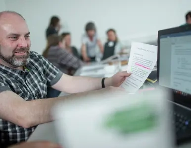 Ein Herr sitzt vor einem Computerbildschirm  und hält ein Blatt Papier neben den Bildschirm. Im Vordergrund ist unscharf eine Broschüre zu sehen. Im Hintergrund sind unscharf weitere Personen zu sehen.
