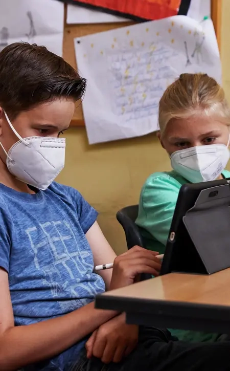Ein Junge und ein Mädchen sitzen an einem Tisch nebeneinander. Sie haben beide ein Tablet vor sich stehen. Sie schauen aber beide auf das Tablet von dem Jungen.
