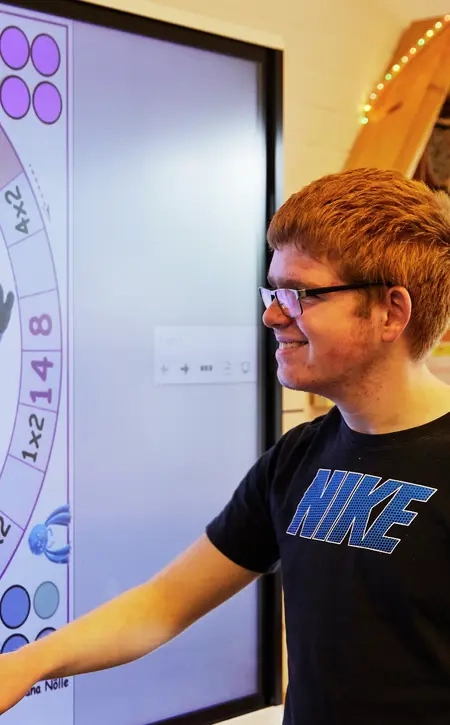 Ein Junge und ein Mädchen stehen vor einem Smartboard und betrachten es. Das Mädchen ist nur von hinten zu sehen, der Junge von der Seite.