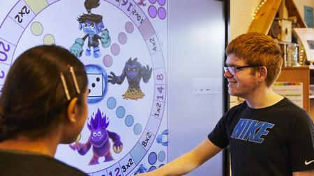 Ein Junge und ein Mädchen stehen vor einem Smartboard und betrachten es. Das Mädchen ist nur von hinten zu sehen, der Junge von der Seite.