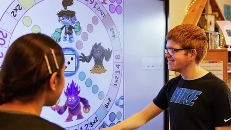 Ein Junge und ein Mädchen stehen vor einem Smartboard und betrachten es. Das Mädchen ist nur von hinten zu sehen, der Junge von der Seite.