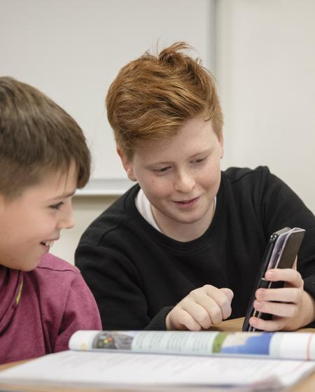 Zwei Jungen sitzen an einem Schreibtisch und schauen gemeinsam auf ein Handy.
