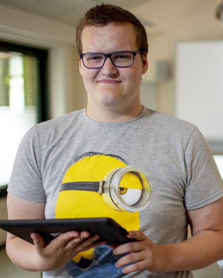 Eine Junge hält ein Tablet in der Hand und lacht in die Kamera. Im Hintergrund ist unscharf ein Klassenraum mit Computern zu sehen.