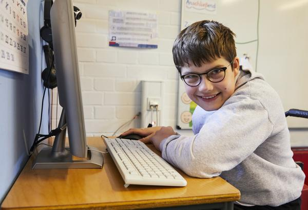 Ein Junge sitzt vor einem Computer. Er ist von der Seite fotografiert und lächelt in die Kamera.