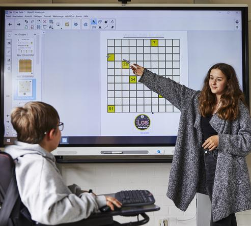 Eine junge Frau zeigt etwas auf einem Smartboard. Ein Kind im  Rollstuhl schaut zu ihr hin.