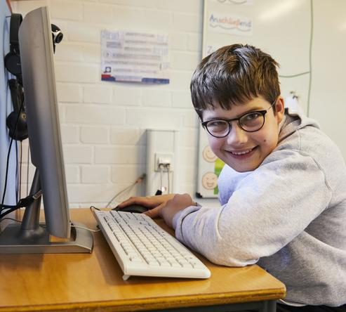Ein Junge sitzt vor einem Computer. Er ist von der Seite fotografiert und lächelt in die Kamera.
