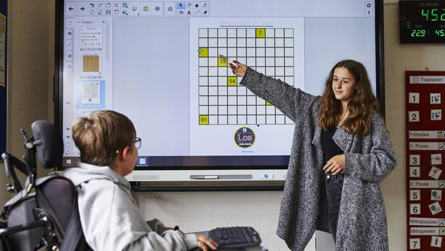 Eine junge Frau zeigt etwas auf einem Smartboard. Ein Kind im  Rollstuhl schaut zu ihr hin.