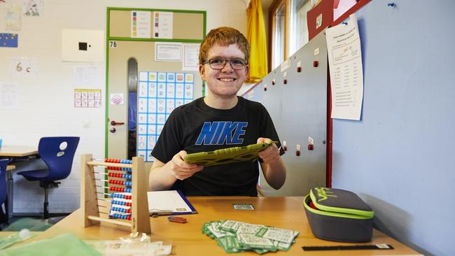 Ein Junge sitzt lächelnd vor einem Tisch. In der Hand hält er ein iPad. Auf dem Tische sind ein Rechenrahmen, karten, ein Mäppchen sowie Papier.