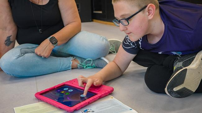 Ein Junge sitzt auf dem Boden und berührt mit einem Finger ein Tablet, das vor ihm liegt. Im Anschnitt ist eine weitere Person zu sehen, die neben ihm auf dem Boden sitzt.