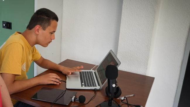 Ein junger Mann sitzt an einem Tisch auf dem sich ein Laptop, ein Tablet sowie ein Mikrofon befinden.