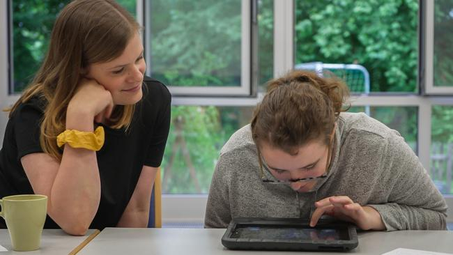 Eine junge Frau sitzt an einem Tisch und schaut auf ein Tablet. Eine andere junge Frau sitzt neben ihr und hat ihren Blick auf das Tablet gerichtet.