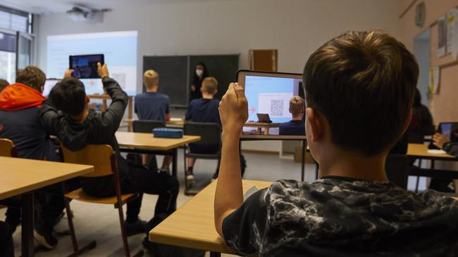 Ein Klassenraum von hinten. Zwei Schüler halten ihre Tablets hoch und fotografieren den QR-Code, der vorne an der Wand zu sehen ist.