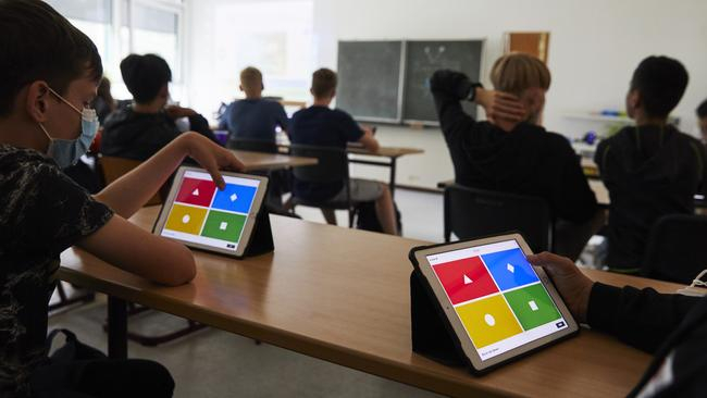Ein Klassenraum von hinten. Vorne sitzen Schüler*innen. Vor den Kindern in der letzten Reihe steht jeweils ein Tablet, das vier farbige Flächen zeigt. Beide Kinder berühren das Tablet an der Stelle mit der blauen Fläche.