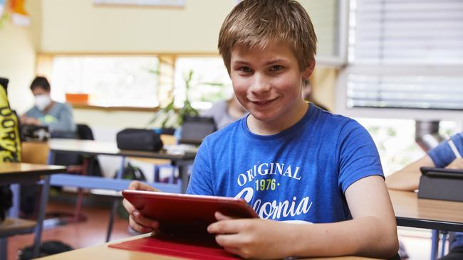 Ein Junge sitzt im Klassenraum an einem Tisch und schaut lächelnd in die Kamera. In der Hand hält er ein Tablet.