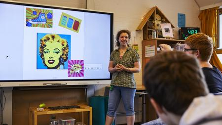 Eine Lehrerin steht vor einem Smartboard. Zwei Schüler schauen zu ihr hin.