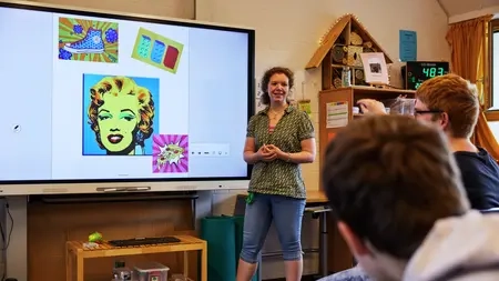 Eine Lehrerin steht vor einem Smartboard. Zwei Schüler schauen zu ihr hin.