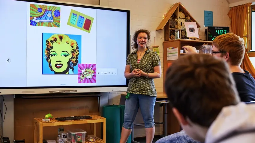 Eine Lehrerin steht vor einem Smartboard. Zwei Schüler schauen zu ihr hin.