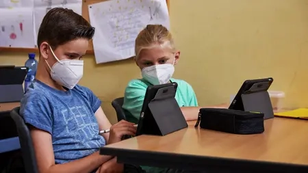 Ein Junge und ein Mädchen sitzen an einem Tisch nebeneinander. Sie haben beide ein Tablet vor sich stehen. Sie schauen aber beide auf das Tablet von dem Jungen.