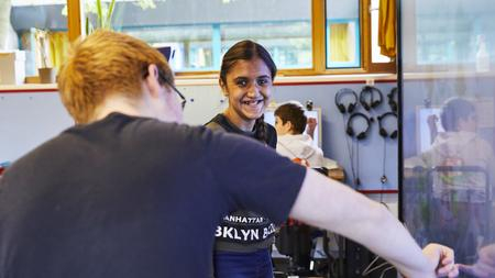 Zwei Jugendliche stehen vor einem Smartboard. Den Jungen sieht man nur von hinten. Das Mädchen sieht man von vorne, es lacht.