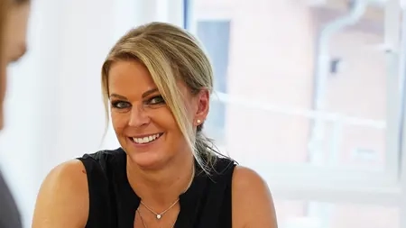 Silke Müller, eine Frau mit blonden Haaren, lächelt in die Kamera. Im Vordergrund sieht man unscharf eine weitere Person sowie einen Laptop, der vor ihr auf dem Tisch steht.