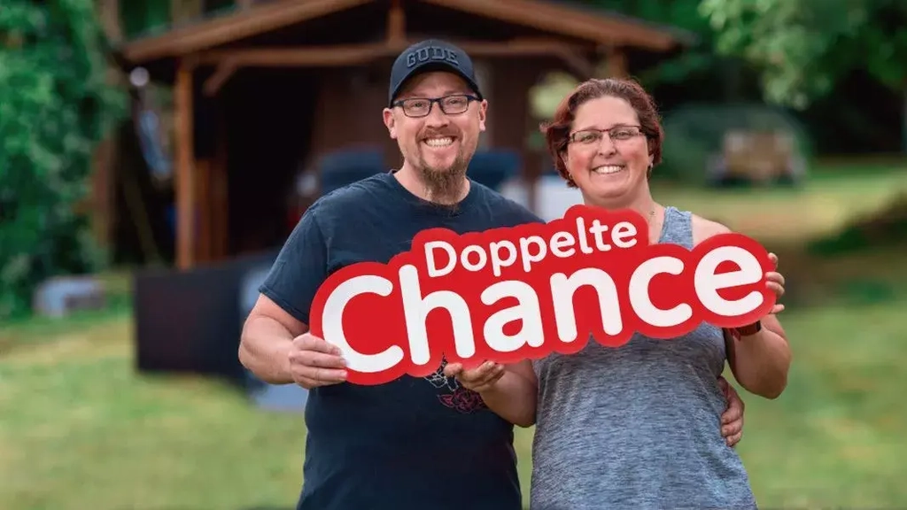 Aktion Mensch-Gewinnern Sandra steht lachend neben ihrem Mann im gemeinsamen Garten. Sie halten den Schriftzug "Doppelte Chance" in der Hand.