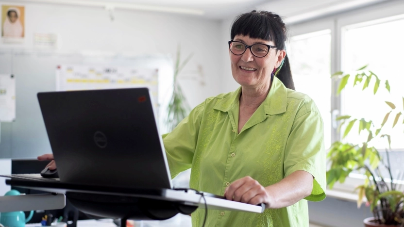 Eine Frau steht in einem Büroraum an einem Laptop. Sie trägt einen Brille, einen Pferdeschwanz und eine hellgrüne, kurzärmelige Bluse.