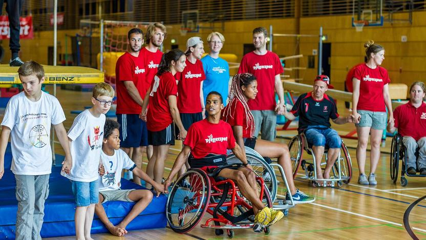 Eine Gruppe Menschen hält sich in einer Reihe an den Händen. Manche sitzen im Rollstuhl. Viele tragen ein rotes T-Shirt mit der Aufschrift "Pfeffersport".