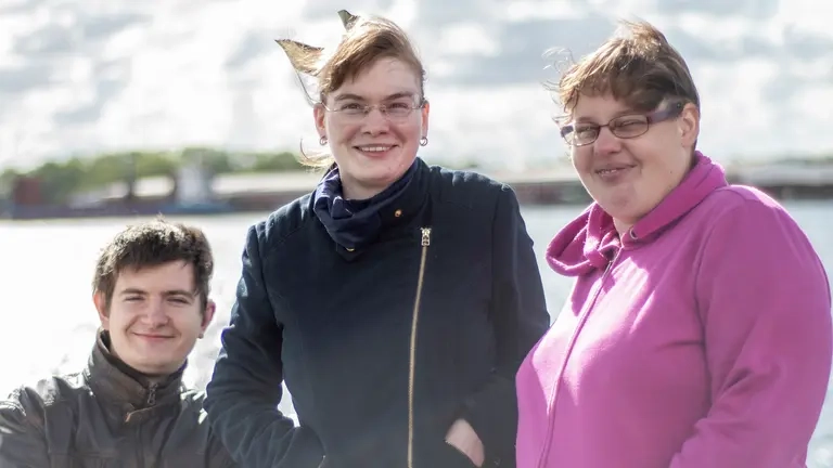 Drei Personen stehen an einem Geländer, hinter ihnen ist das Meer zu sehen, sie sind warm angezogen und es scheint windig zu sein.