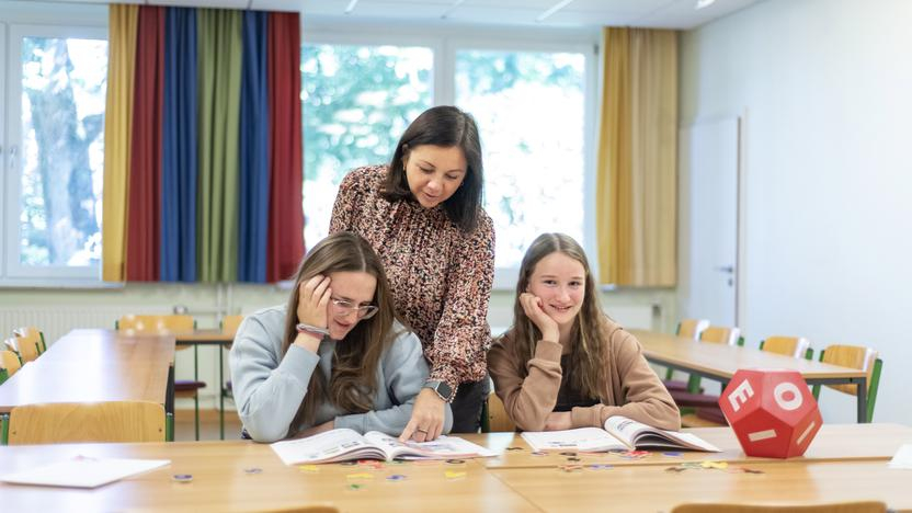Zwei Mädchen lesen in einem Buch, eine Frau zeigt im Buch auf etwas.