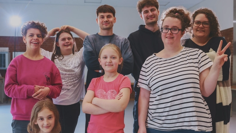 Eine achtköpfige Gruppe von Jungen und Mädchen, in der Altersspanne von etwa 10 bis ca. 25 posieren zusammen in einem Tanzstudio für das Foto.