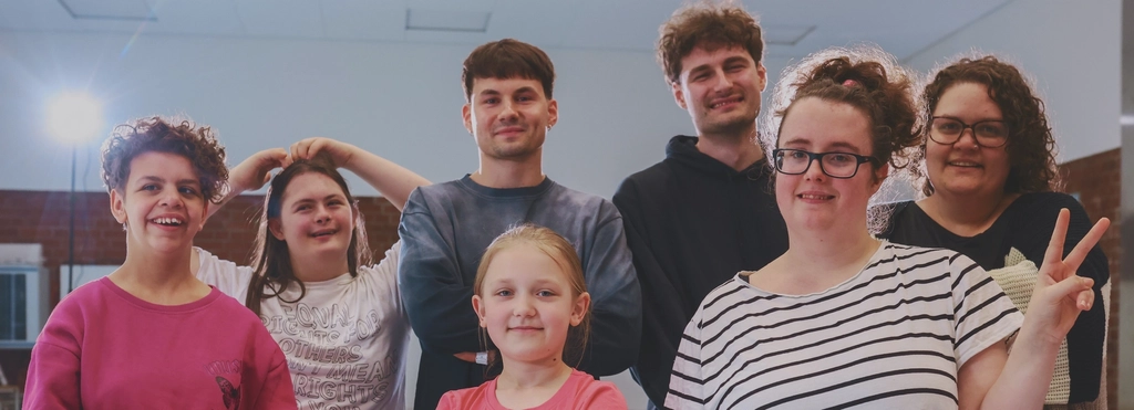 Eine achtköpfige Gruppe von Jungen und Mädchen, in der Altersspanne von etwa 10 bis ca. 25 posieren zusammen in einem Tanzstudio für das Foto.