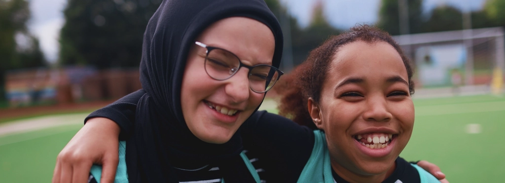 Zwei Mädchen, eines davon mit Kopftuch, stehen lachend Arm in Arm auf einem Sportplatz.