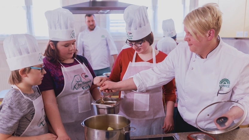 Vor einem großen Topf auf dem Herd stehen zwei größere Mädchen und ein kleinerer Junge mit Kochmütze und Schürzen. Eine junge Frau mit Kochjacke und kurzen blonden Haaren neben ihnen hilft ihnen, eine Soße anzurühren.