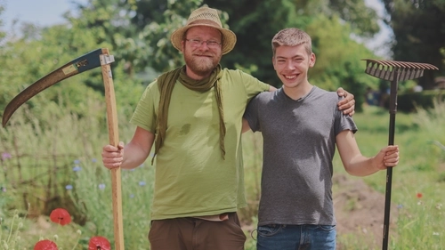 Ein Mann mit Hut und ein Jugendlicher haben einen Arm um die Schulter des anderen gelegt. In der freien Hand halten si eine Sense und einen Rechen. Sie lachen in die Kamera.