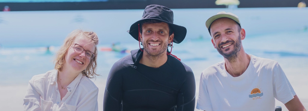 Eine junge Frau und zwei junge Männer sitzen lachend vor einem Schwimmbecken. Zwei von ihnen sind Rollstuhlfahrer*innen.