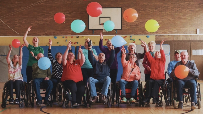 Eine Gruppe Rollstuhlfahrer*innen mit Ballons in einer Turnhalle