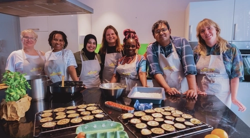 Sieben personen stehen lächelnd hinter einer Arbeitsplatte mit Speisen in einer Küche