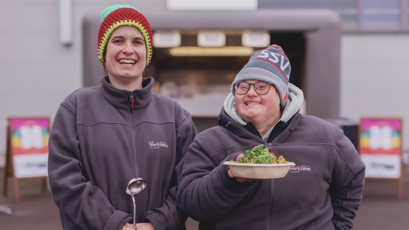 Zwei Frauen posieren vor einem Foodtruck-Anhänger, der etwas verschwommen im Hintergrund zu sehen ist, und lachen. Beide tragen Mützen und einheitliche graue Jacken. Die Frau rechts im Bild hat eine sichtbare Behinderung. Sie hält stolz einen Teller mit Essen hoch, als ob sie ihn anbieten würde. Die Frau neben ihr hält eine Suppenkelle in den Händen.