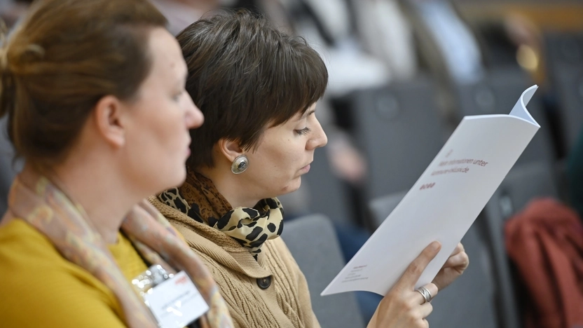 Eine Frau liest in einer Broschüre, neben ihr sitzt eine weitere Frau und schaut mit hinein