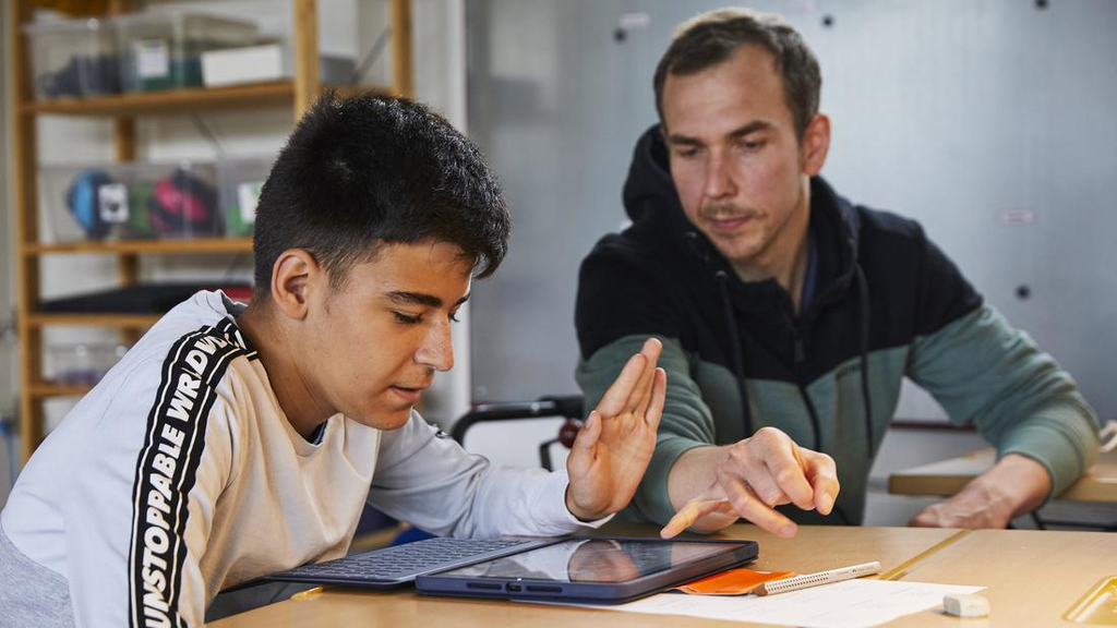 Ein Jugendlicher sitzt an einem Tisch und schaut auf ein Tablet. Neben ihm sitzt Fabio Priano und weist auf etwas auf dem Tablet.