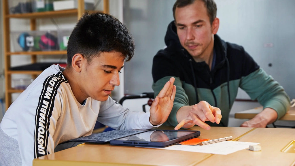 Ein Jugendlicher sitzt an einem Tisch und schaut auf ein Tablet. Neben ihm sitzt Fabio Priano und weist auf etwas auf dem Tablet.
