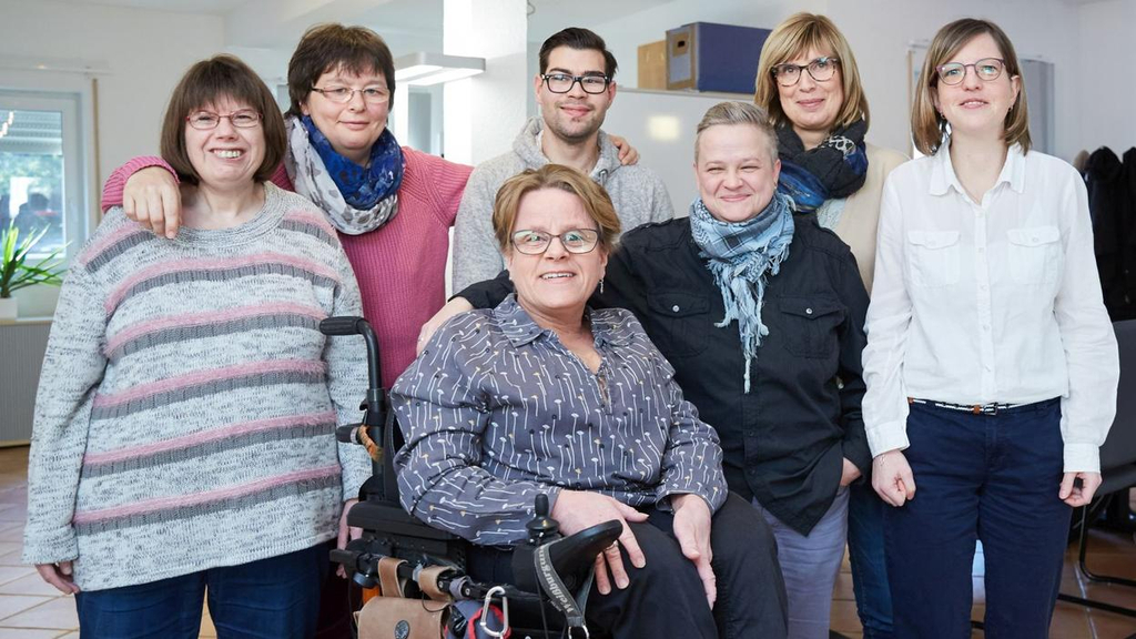 Sechs Frauen und ein Mann stehen für Gruppenbild zusammen. Die Frau in der Mitte sitzt in einem E-Rolli.