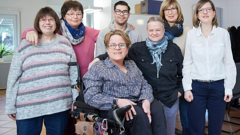 Sechs Frauen und ein Mann stehen für Gruppenbild zusammen. Die Frau in der Mitte sitzt in einem E-Rolli.