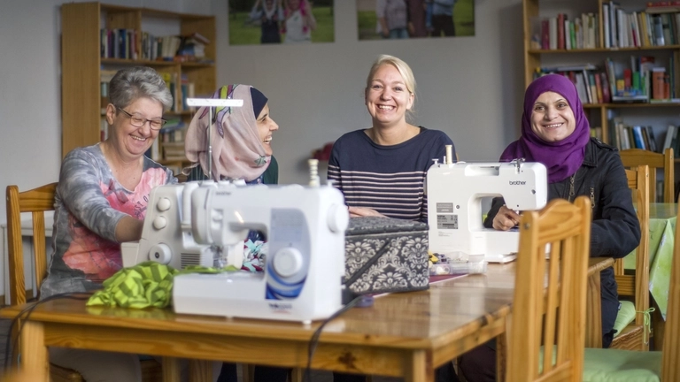 Vier Frauen sitzen an Nähmaschinen, lachen und nähen gemeinsam.